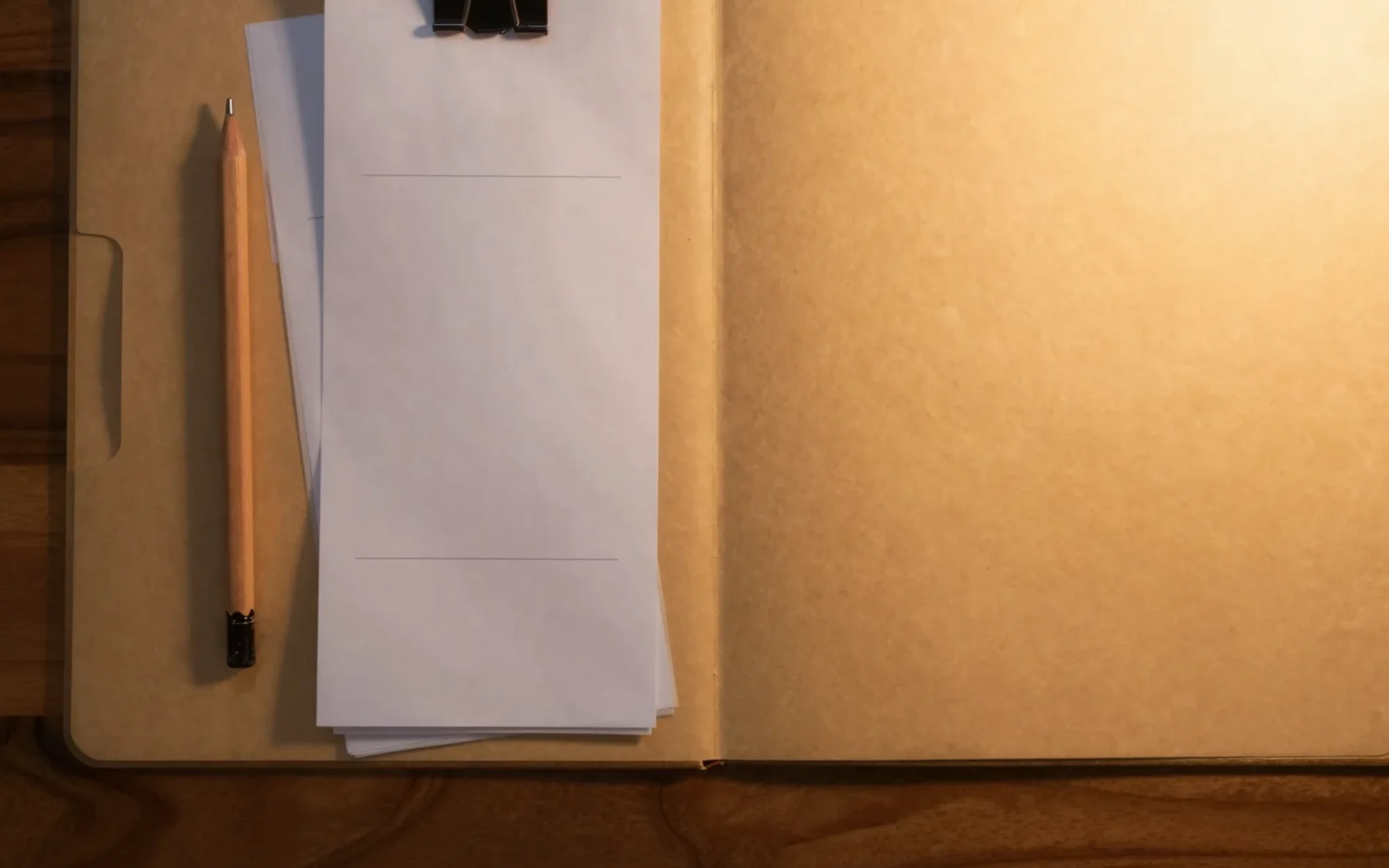 An open tan folder with blank receipts held by a binder clip, a pencil beside it, on a wooden table in lamp light